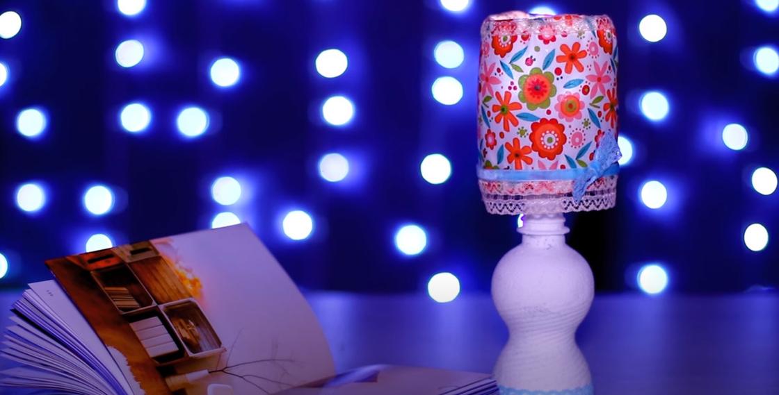 На столе стоит лампа с цветным абажуром и белой пластиковой ножкой. Рядом лежит раскрытая книга