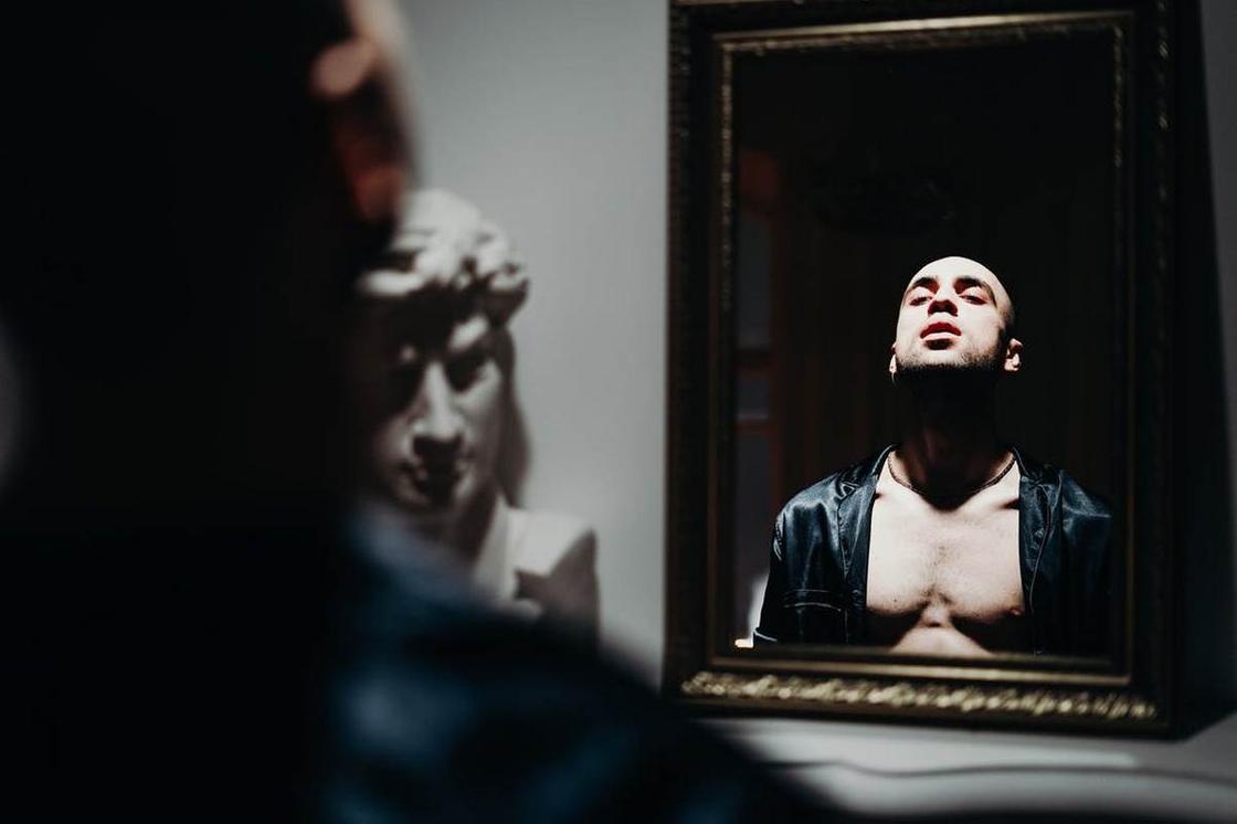 Мужчина в расстегнутой черной рубашке смотрит на свое отражение в зеркале