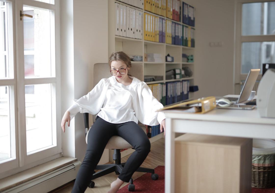 Девушка в белой рубашке и темных брюках сидит, развалившись в кресле, возле рабочего стола