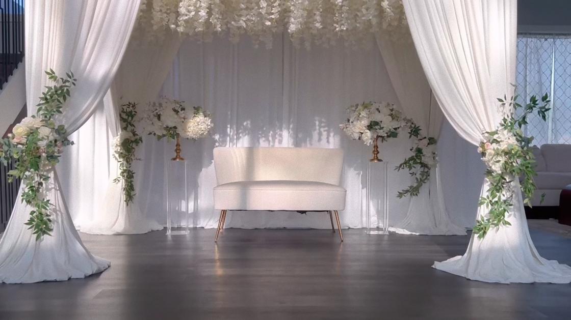Фотозона в свадебном зале сделана в виде беседки с балдахином. По центру стоит мини-диван, все украшено белыми искусственными цветами с зеленью