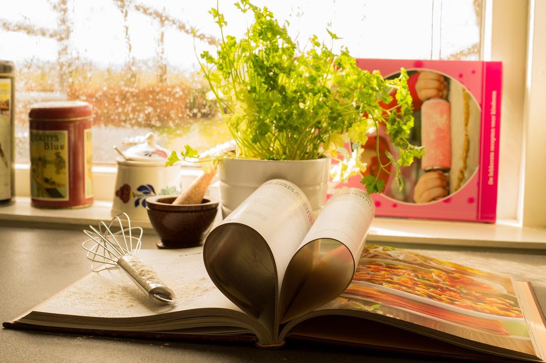 Книга с рецептами лежит на рабочем столе рядом с венчиком и ступкой с пестиками