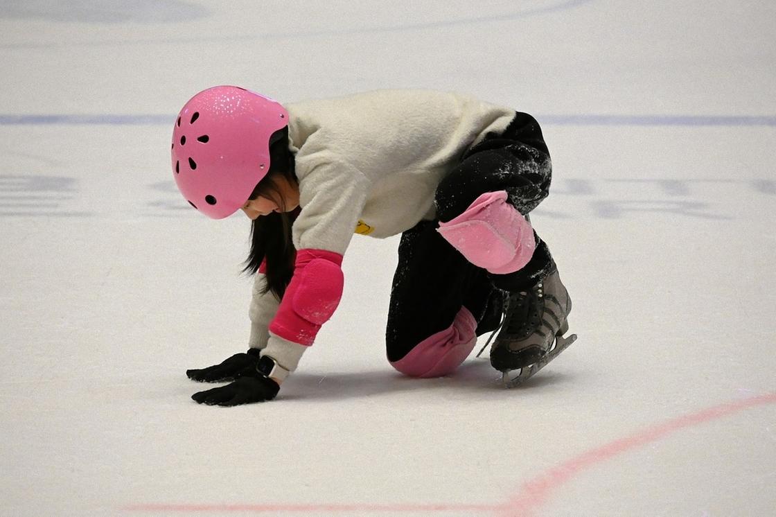 Девочка в экипировке на коньках упала на льду на колено и уперлась руками в лед