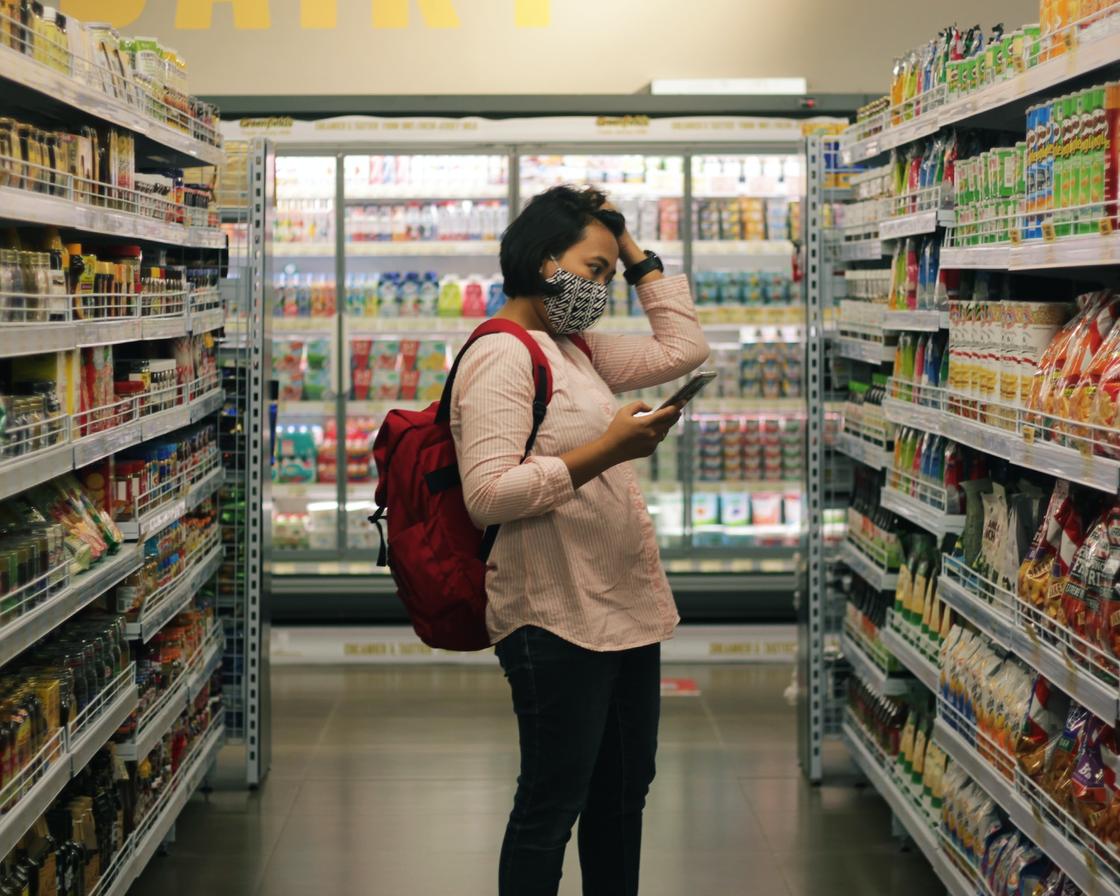 Женщина с рюкзаком стоит среди полок в магазине, держит в руках телефон