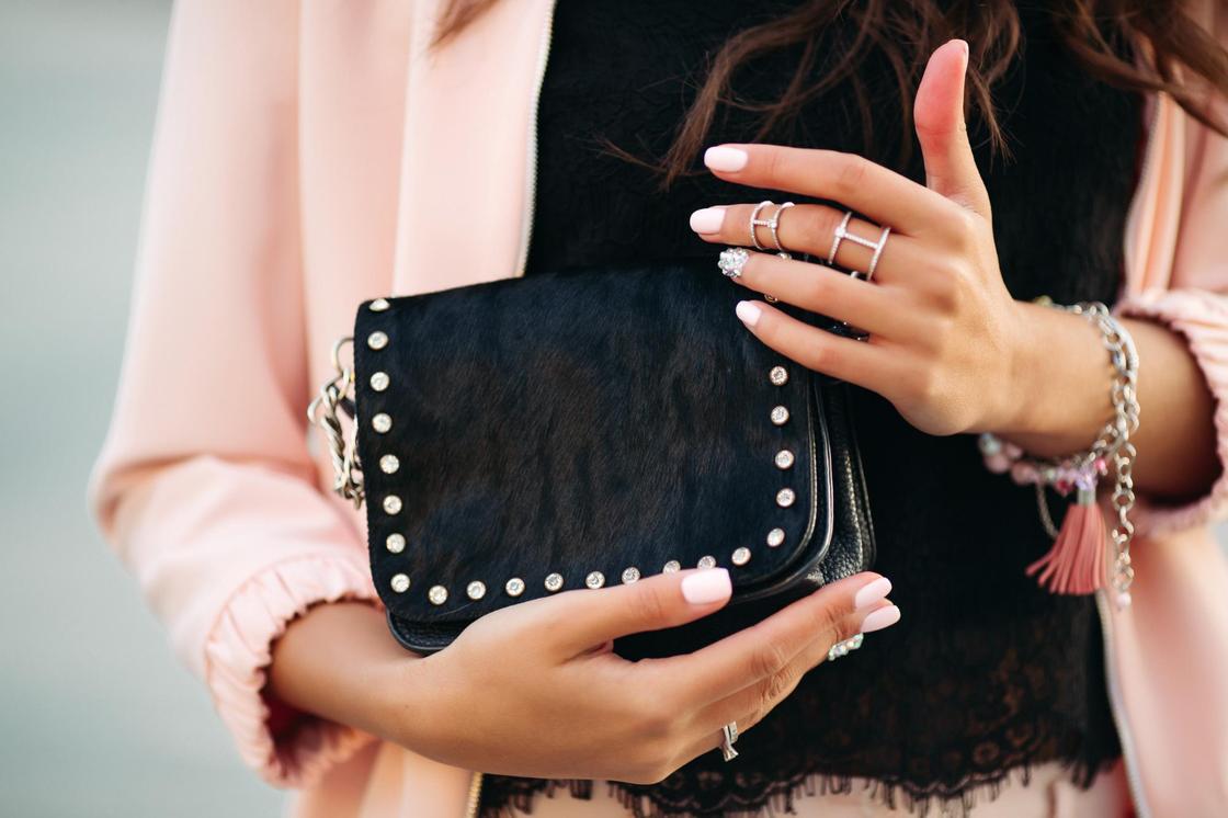 Девушка в кружевной блузе и розовой куртке держит в руках, украшенных кольцами и браслетом, черную сумочку