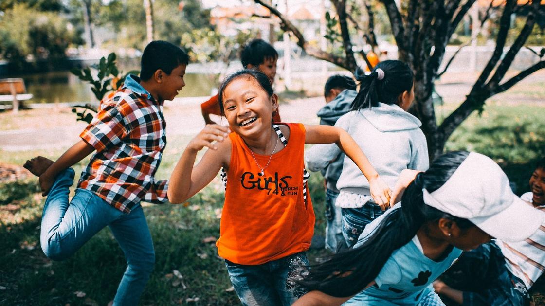 Девочка в оранжевой майке бежит и смеется, мальчик прыгает на одной ноге, а другие дети бегут в разные стороны