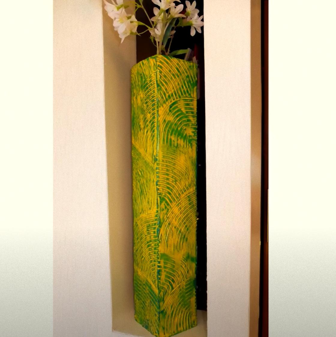 В нише стоит высокая четырехгранная напольная ваза с цветами. Поверхность вазы рельефная и окрашена в желто-зеленый цвет