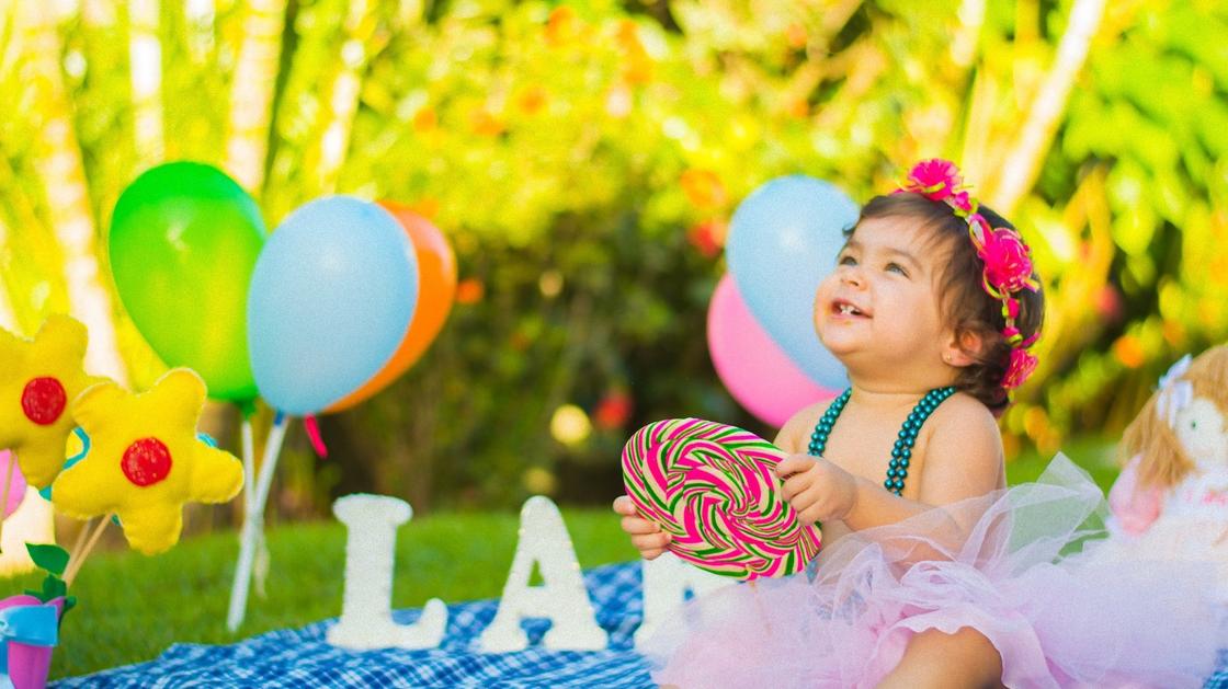 Маленькая девочка в веночке сидит на лужайке с ярким кругом в руках и смеется. Лужайка украшена воздушными шариками, надувными цветами и буквами