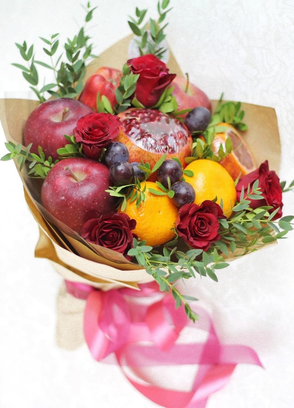 В букете собраны красные яблоки, разрезанный гранат, мандарины, сливы, розы, веточки самшита