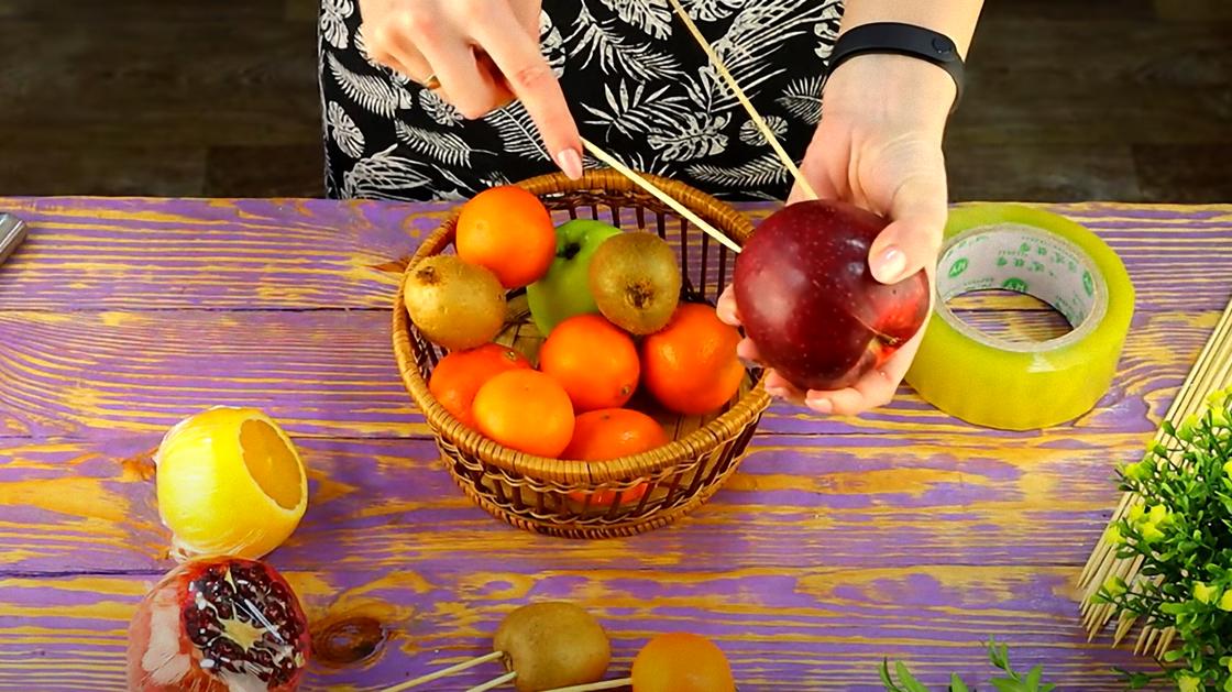 В красное яблоко втыкают деревянные шпажки. На столе стоит корзина с разными фруктами, лежит апельсин и гранат со срезанными верхушками, киви на шпажках, скотч и шпажки