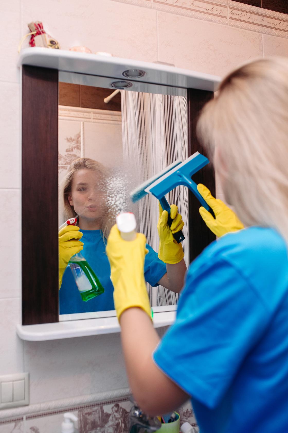 Женщина моет зеркало ракелем. Она опрыскивает поверхность чистящимспреем
