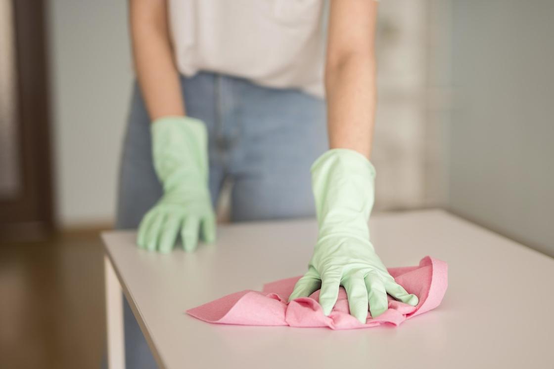 Руками в резиновых перчатках держат розовую салфетку и вытирают пыль на столе