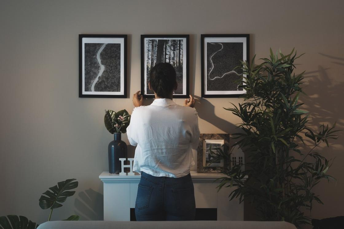 Женщина в белой блузке вешает третью черно-белую картину на стену. Рядом стоит высокое комнатное растение и тумбочка с вазой, картиной, вырубкой