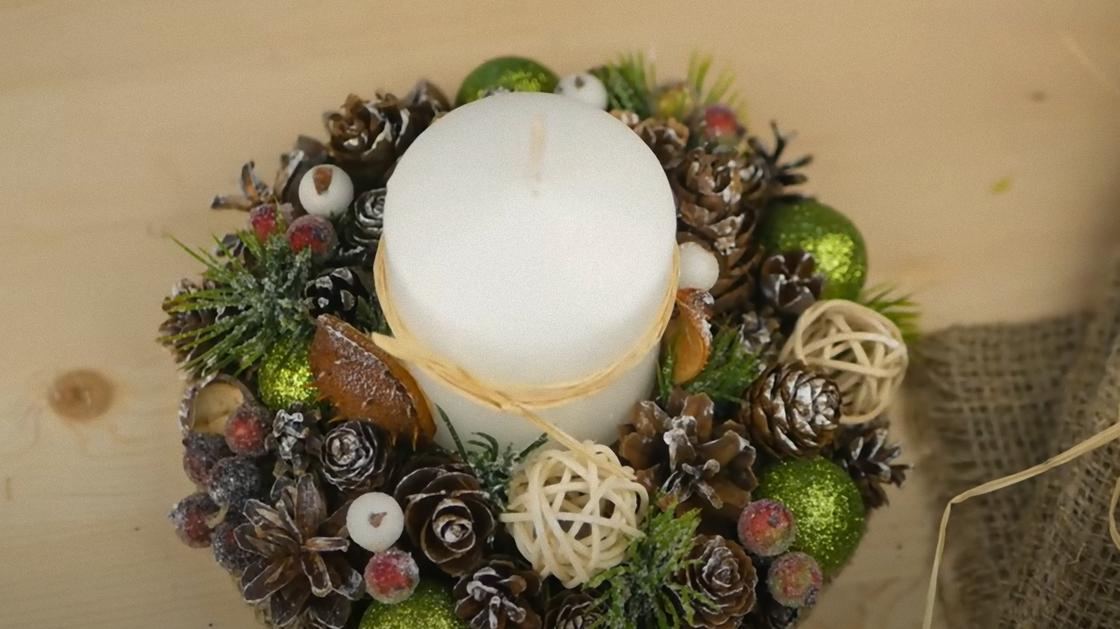 Белая свеча стоит на подсвечнике, украшенном шишками, хвоей, шариками, искусственными ягодами