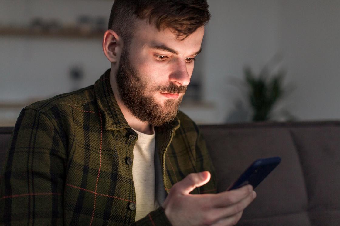 Бородатый мужчина в клетчатой рубашке смотрит в экран телефона