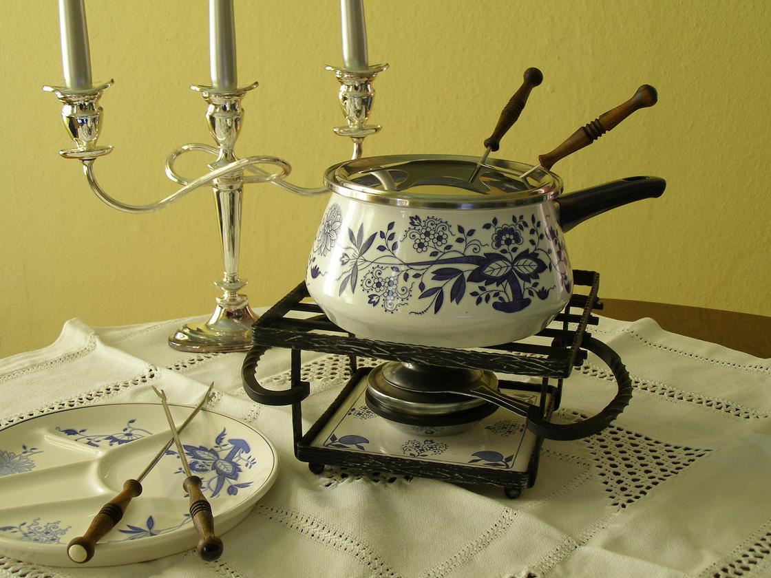 На столе с белой скатертью стоит набор посуды для приготовления фондю: какелон, тарелка, вилки