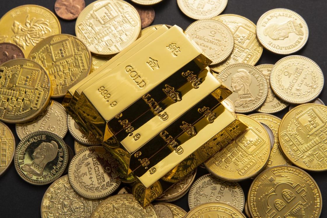 На столе рассыпаны золотые монеты разных номиналов. По центру горкой лежат золотые слитки 999 пробы