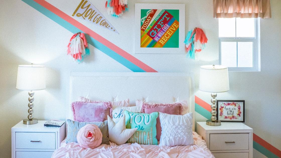 В комнате стоит большая кровать с декоративными подушками, мягкими игрушками. Рядом две прикровтаные тумбочки с настольными лампами, а на стене нарисована разноцветная полоса, висит картина с надписями