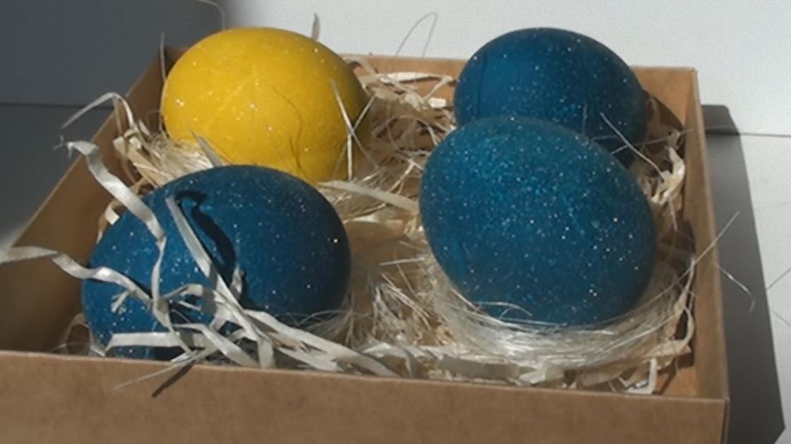 В коробке со стружкой лежат открашенные в синий и желтый цвет яйца. Поверхность яиц с блеском