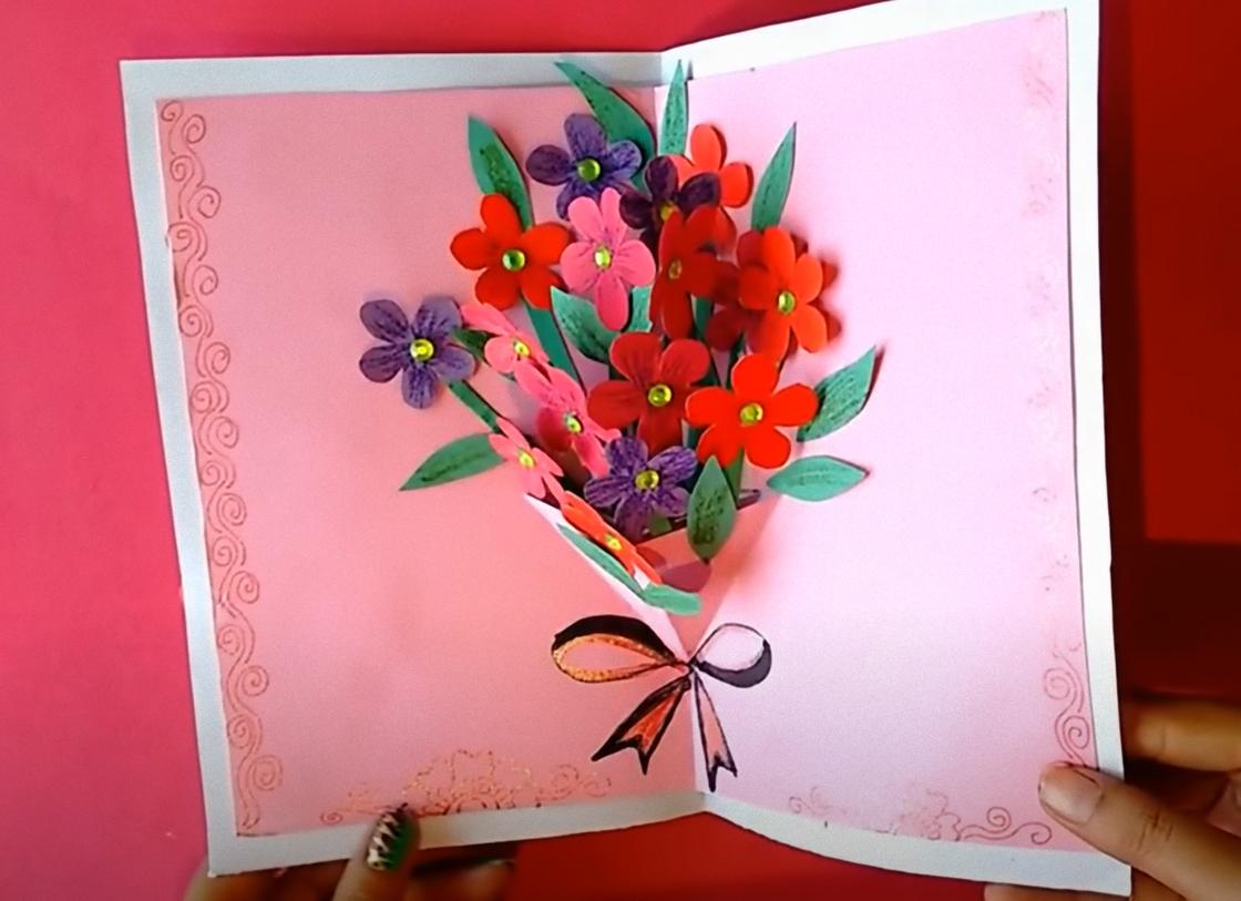 В руках держат открытую открытку с цветочным объемным букетом внутри. Цветочки красного, розового, фиолетового цвета размещены на зеленых стеблях и украшены по центру полубусинами