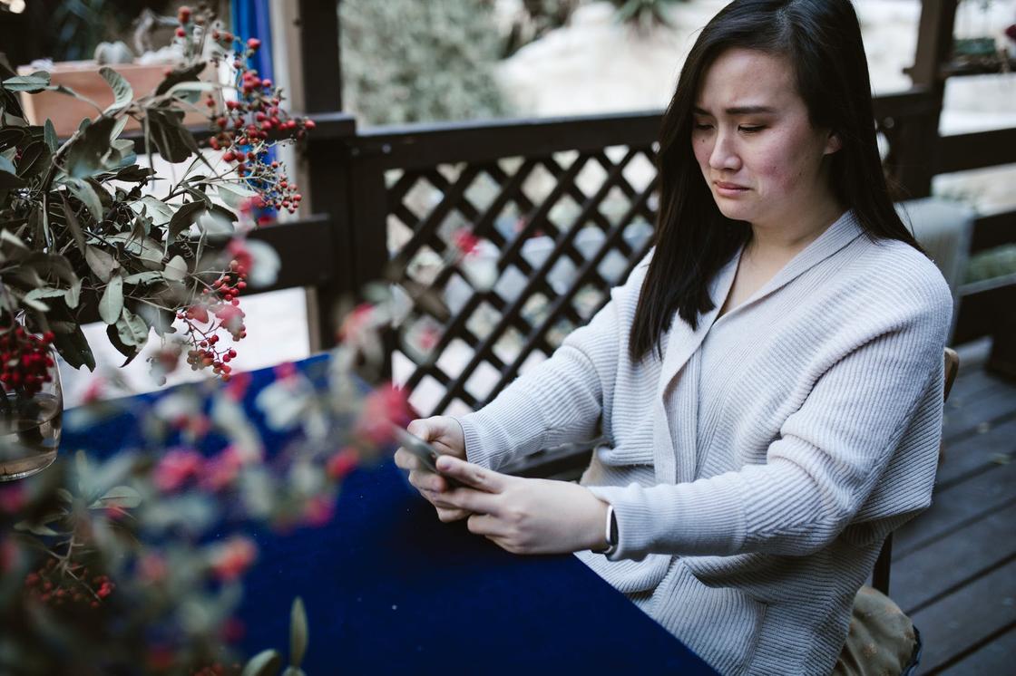 Плачущая девушка сидит за столом на открытой террасе и смотрит на экран телефона