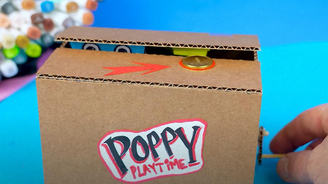 Коробка из картона с надписью «Poppy Playtime» и слегка открытм верхом, откуда выглядывают глаза и тянется рука. Сверху на коробке нарисована красная стрелка и кружок, на котором лежит монета, рукой крутят ручку сбоку ящика