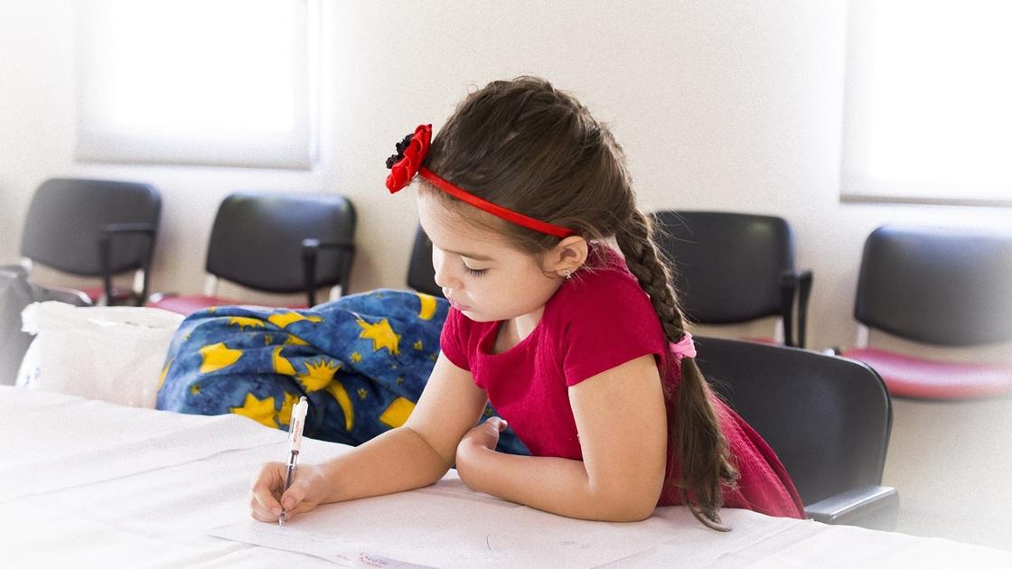 Девочка с длинной косичкой в красной футболке сидит за столом и пишет в тетради