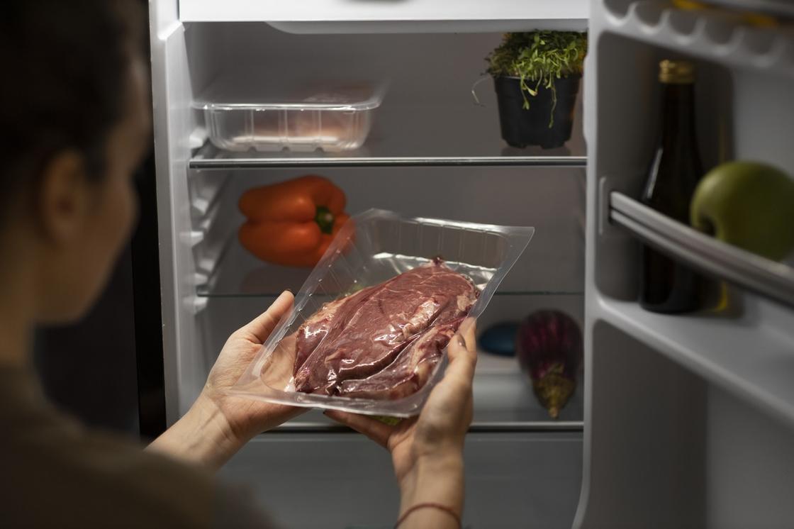 Женщина стоит перед открытым холодильником и держит в руках кусок мяса в пластиковом контейнере. На полках холодильника стоит зелень, лежит сладкий перец и стоит закрытый лоток