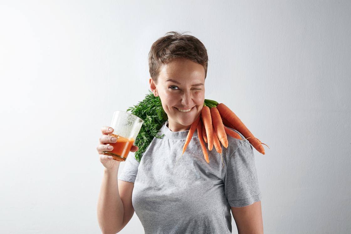 Девушка со связкой моркови на шее держит стакан сока и подмигивает