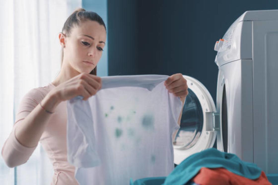 Девушка стоит возле стиральной машины и держит в руках белую блузку с зелеными пятнами