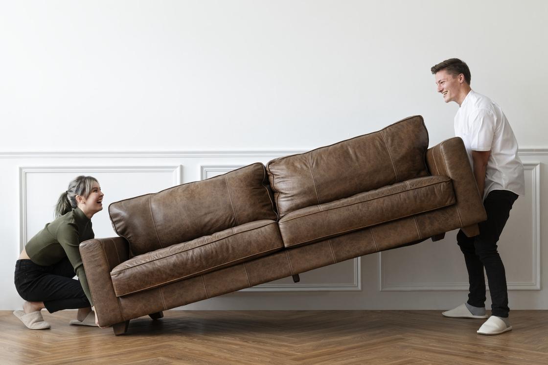 Девушка и парень переставляют кожаный коричневый диван под стенку