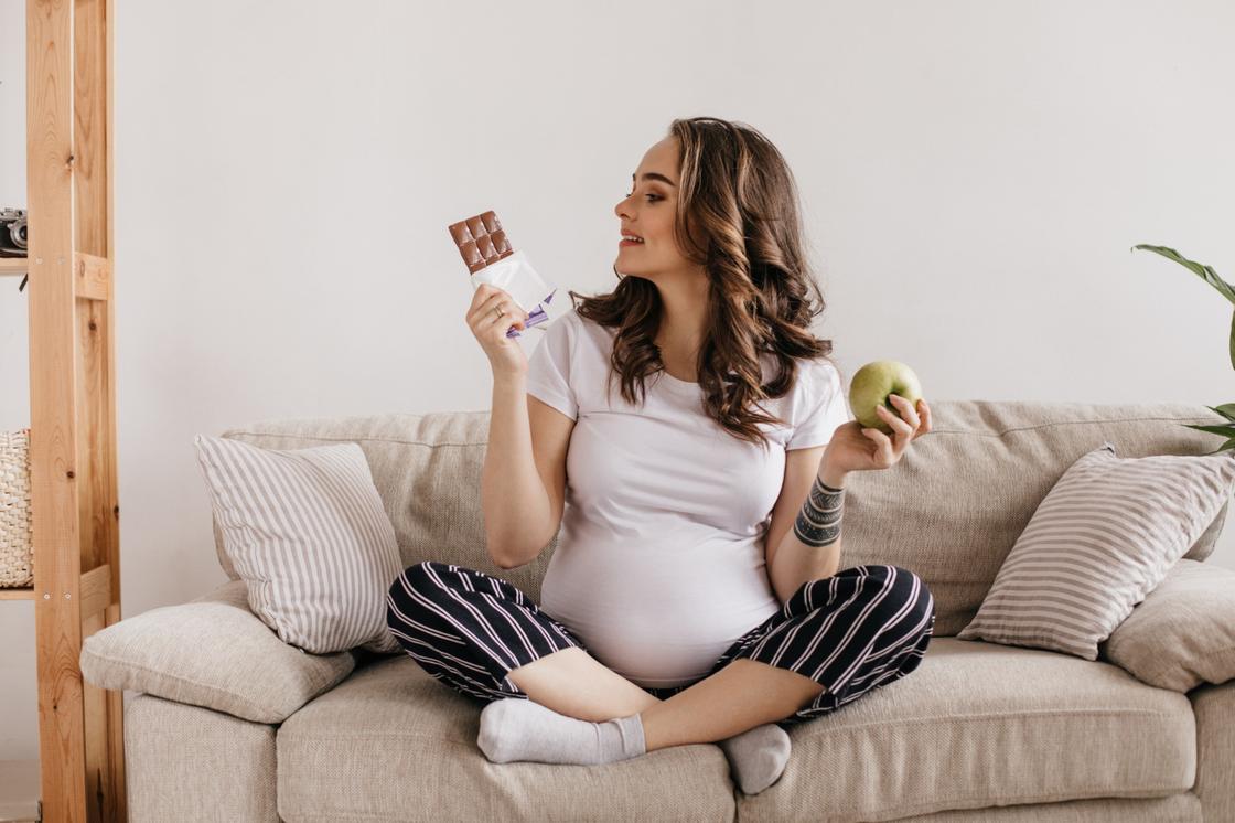 Беременная женщина держит в одной руке яблоко, в другой — плитку шоколада