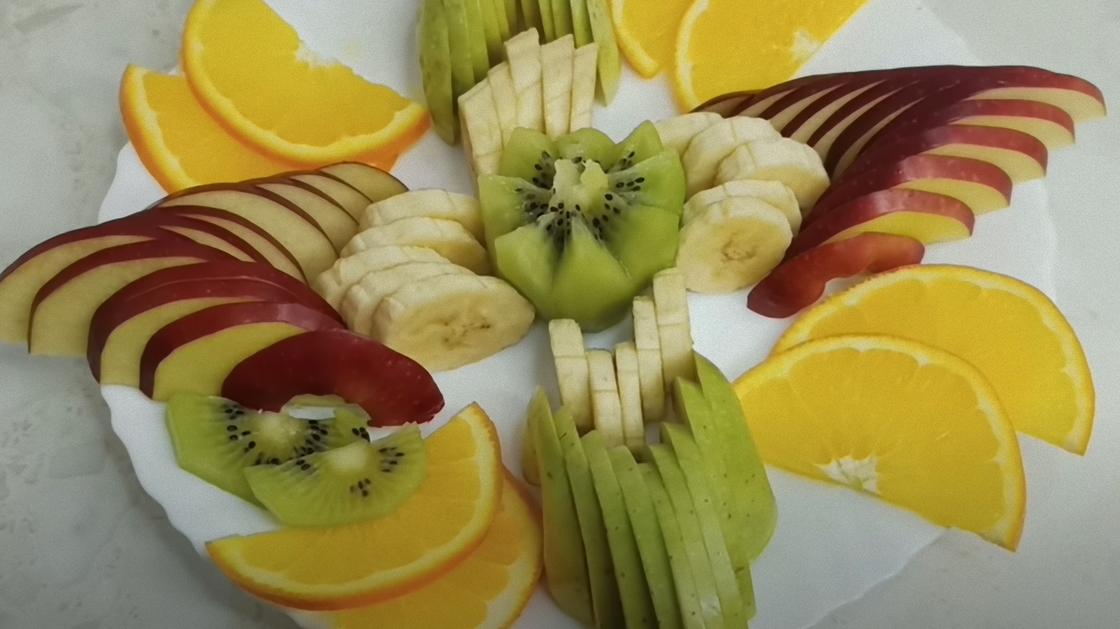 На белой тарелке разложены нарезанные слайсами красные и зеленые яблоки, бананы, апельсины. Киви вырезан в форме цветка и размещен по центру композиции