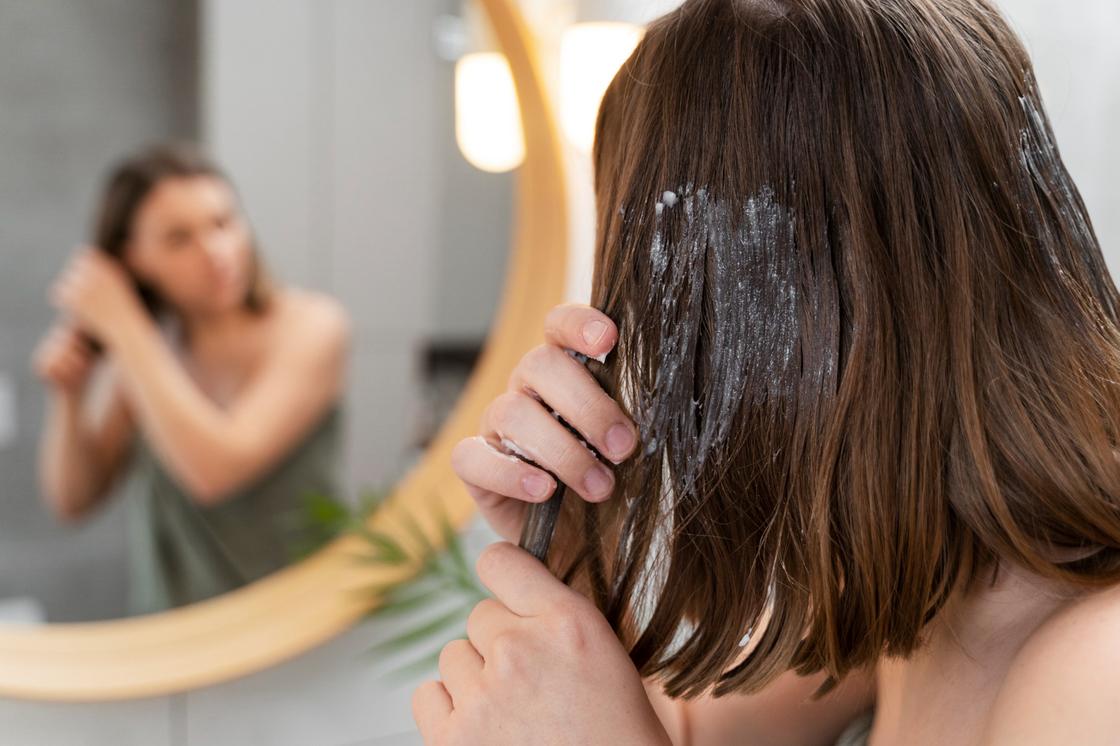 Девушка стоит перед зеркалом и наносит кокосовое масло на волосы