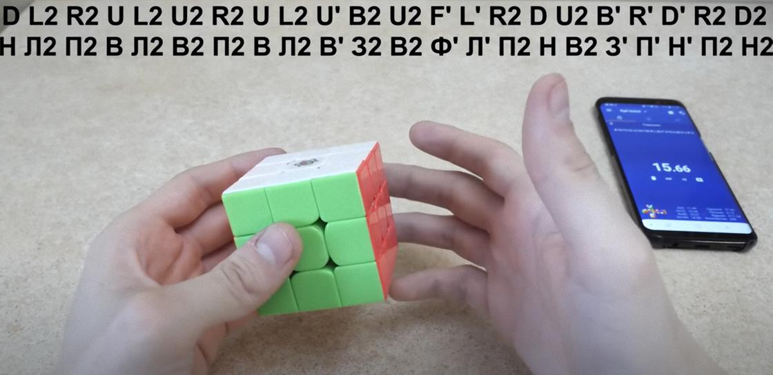 Кубик Рубика в собранном виде держат в руках. Сверху написана формула скрамбла, а рядом лежит телефон