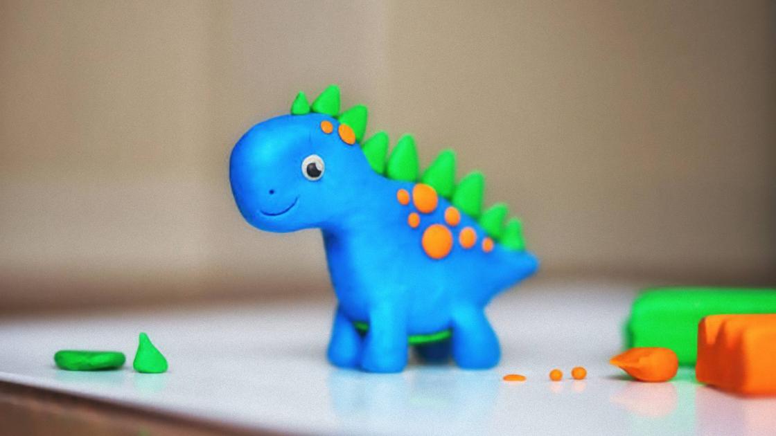 Динозаврик из пластилина стоит на столе, рядом лежат кусочки зеленого и оранжевого пластилина. Туловище динозавра синего цвета, гребень на спине — зеленый, а пятна — оранжевые