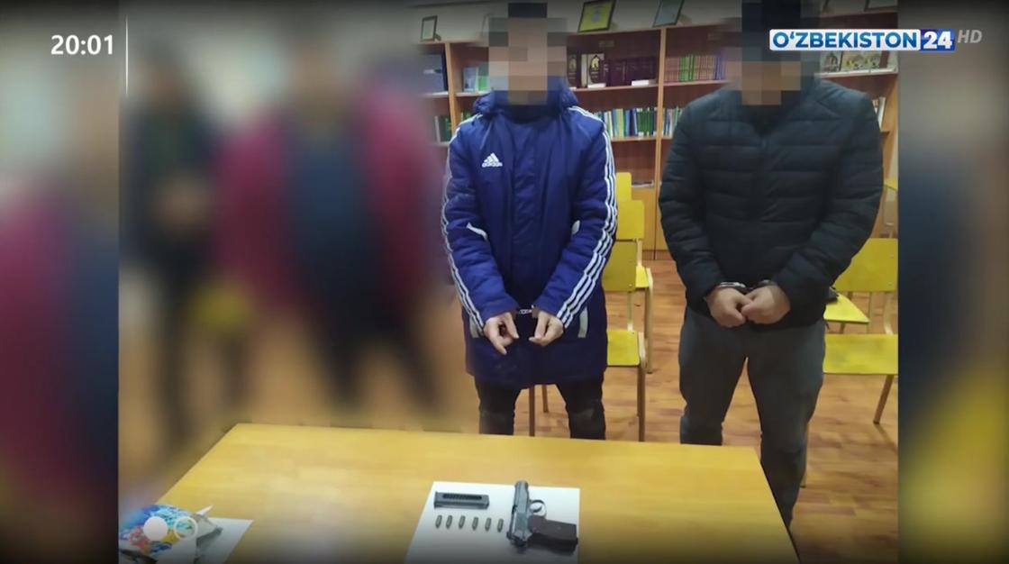 Двое подозреваемых стоят в наручниках перед столом, на котором лежит пистолет