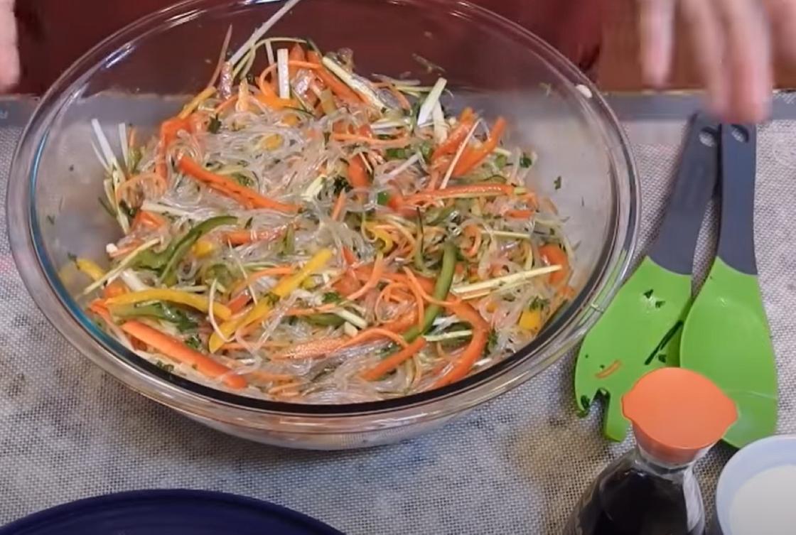 В глубокой стеклянной салатнице нарезанные овощи с лапшой, рядом на столе лежат две зеленые лопатки