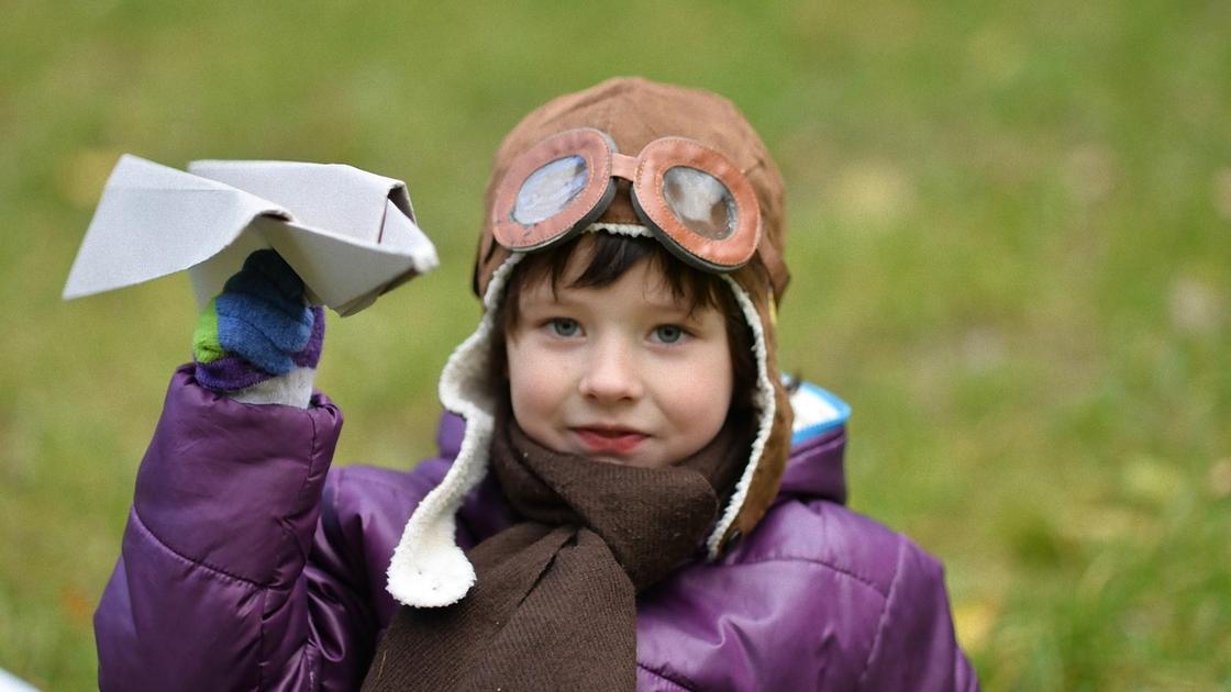 Мальчик одет в шлем летчика держит в руке бумажный самолет