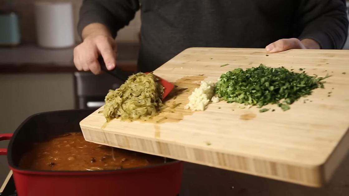 Ингредиенты с разделочной доски: огурцы, зелень, чеснок перекладывают в сковороду с мясом