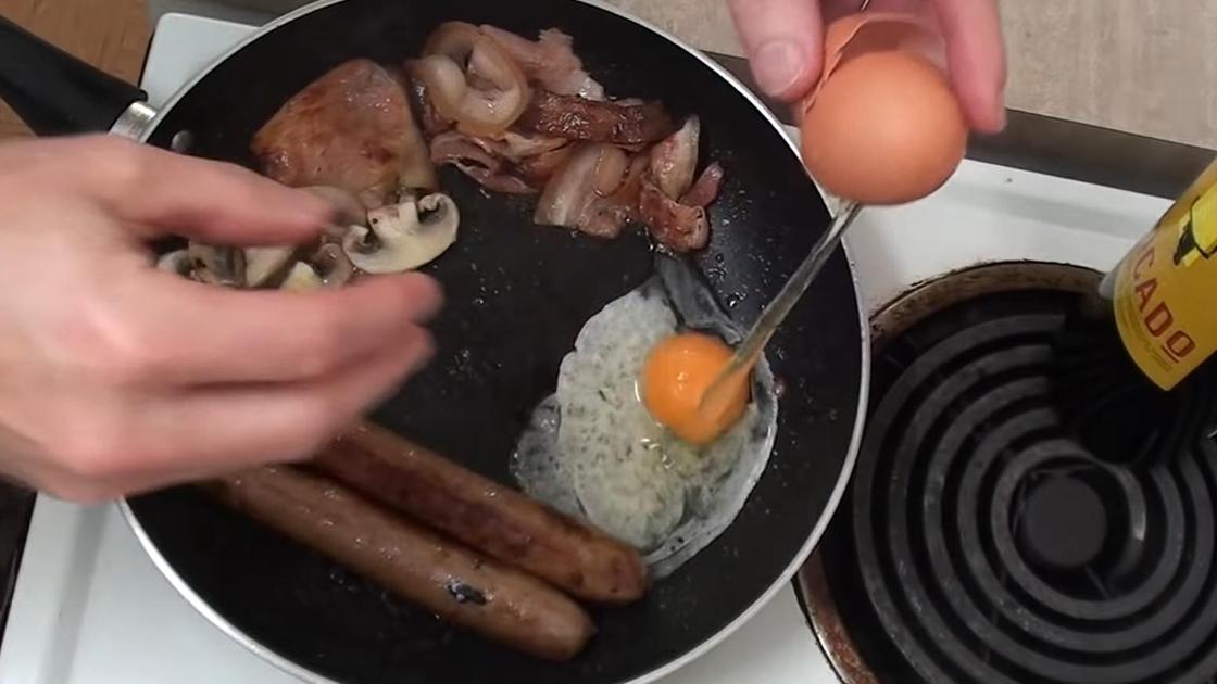 На сковороду с сосисками и беконом разбивают яйцо