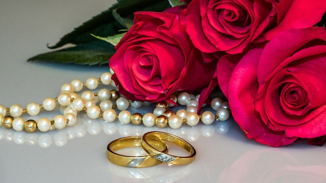 Возле букета красных роз лежат обручальные кольца и жемчужные бусы