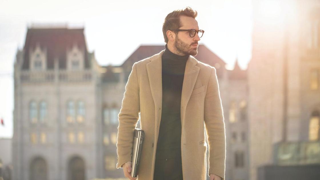 Мужчина в стильном коричневом пальто и очках гуляет по улицам города