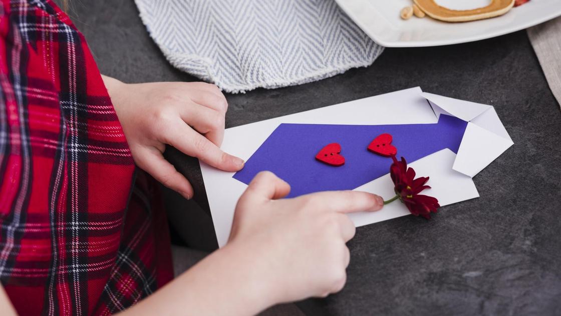 Девочка кладет на стол открытку в виде рбелой рубашки с фиолетовым галстуком. Галстук украшен двумя красными сердечками