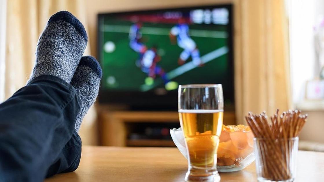 человек закинул ноги на сто, смотрит футбол, на столе пиво и снэки