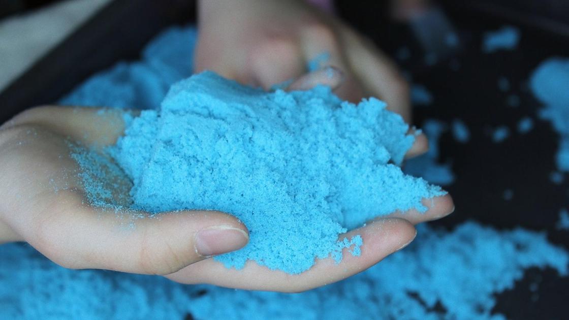 В руке держат горсть голубого кинетического песка. Песок рассыпан на столе