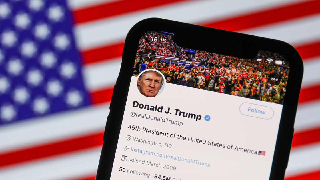 Смартфон с открытой страницей Дональда Трампа в Twitter на фоне государственного флага США