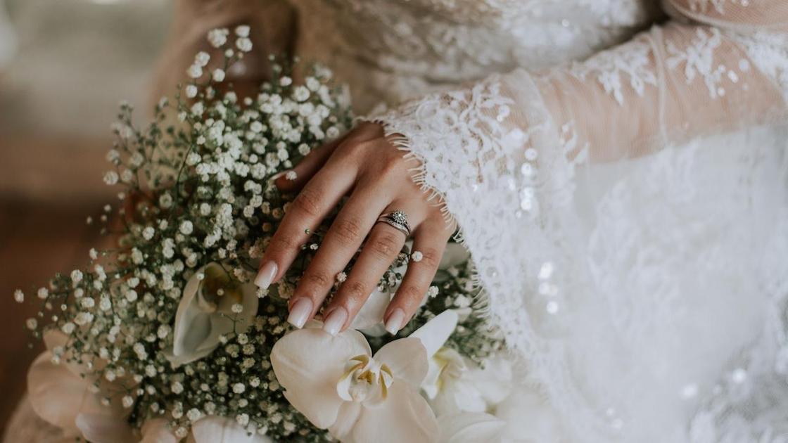 Рука невесты с красивым молочным маникюром и обручальным кольцом лежит на свадебном букете
