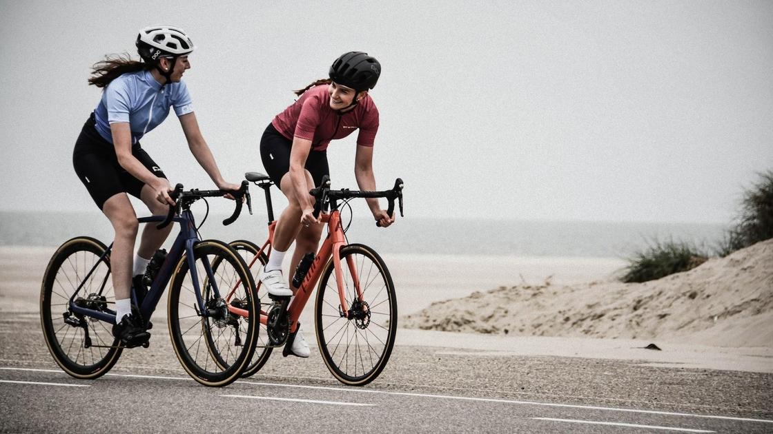 Две девушки в касках едут на спортивных велосипедах по ровной асфальтированной дороге в пустынном месте. Они смотрят друг на друга и улыбаются