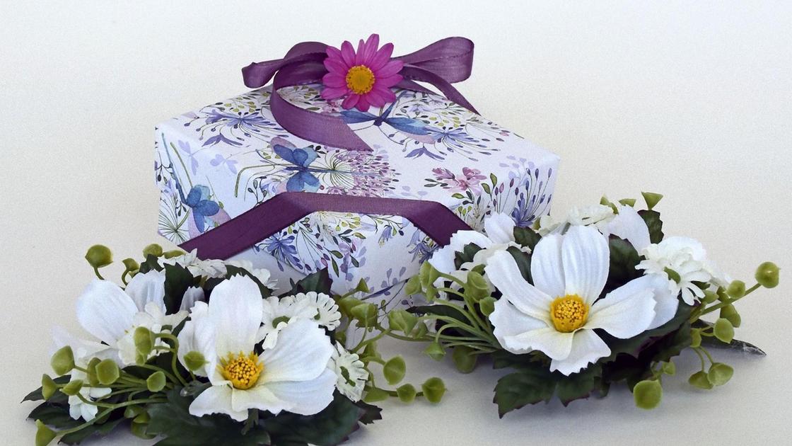 Коробка в красивой упаковочной бумаге с лентой и цветком. Рядом лежит цветочная композиция с белыми цветками