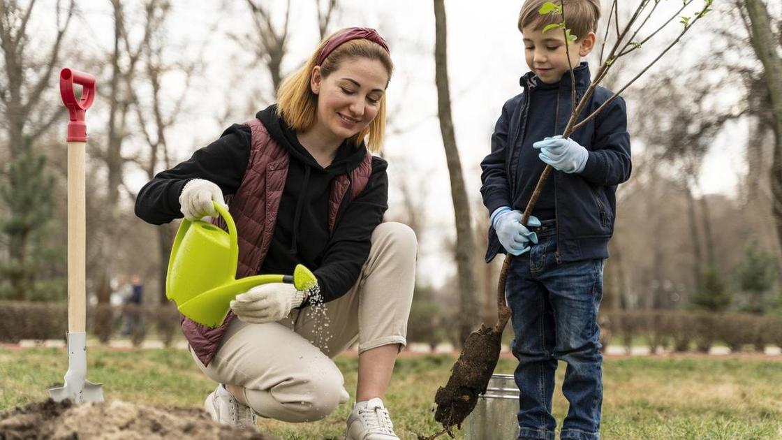Žena nalévá vodu ze zelené konve do díry. Opodál stojí chlapec a v rukou drží sazenici